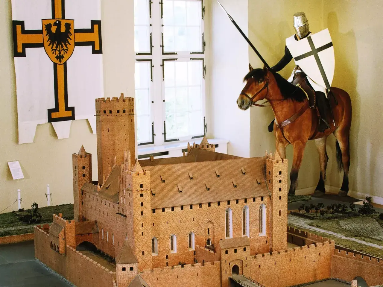 Ein deutscher Orden Ritter auf einem Pferd, mit Lanze und Schild, steht vor einem Modell einer mittelalterlichen Burg.