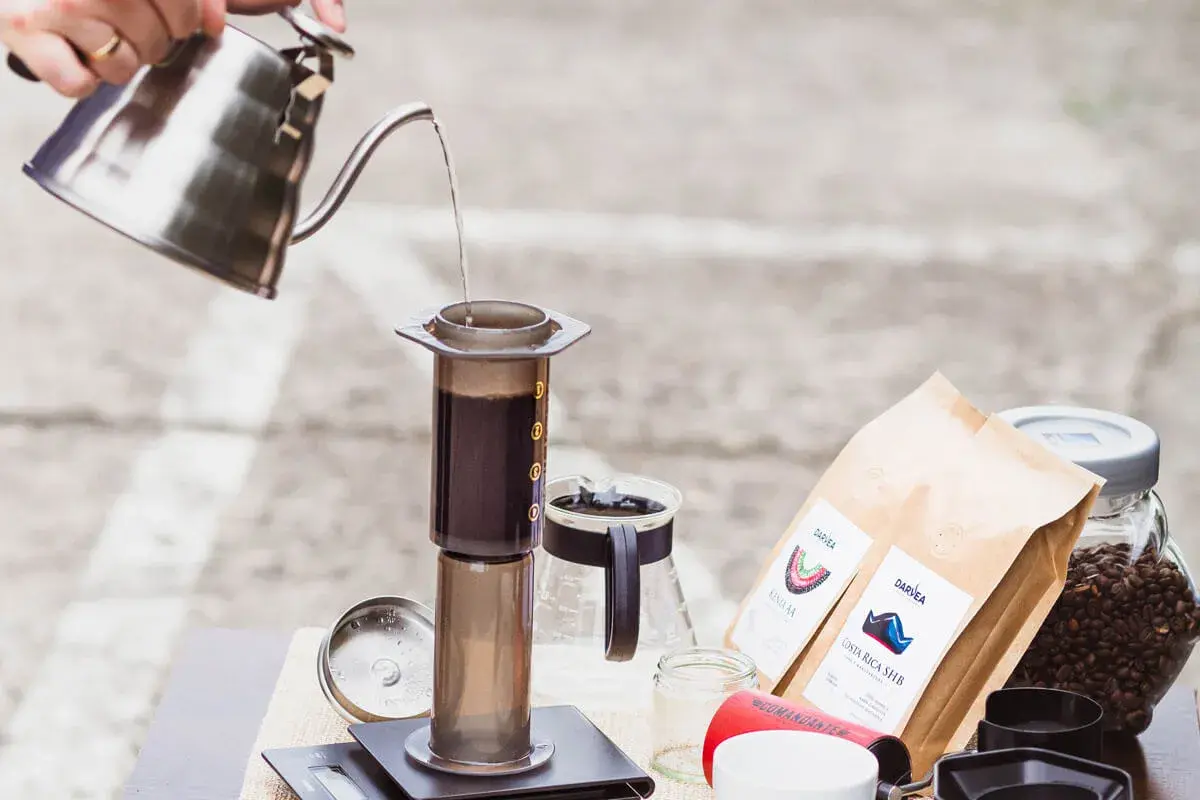 Parzenie kawy metodą Aeropress. Woda nalewana z czajnika do cylindra z kawą. Obok opakowania kawy i ziarna. Czas parzenia jest kluczowy.
