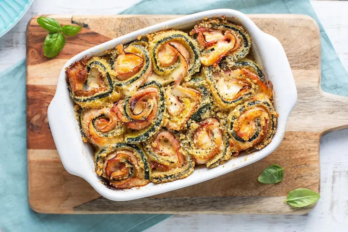 Involtini di zucchine al forno, dorati e filanti, con prosciutto e formaggio, guarniti con basilico fresco.