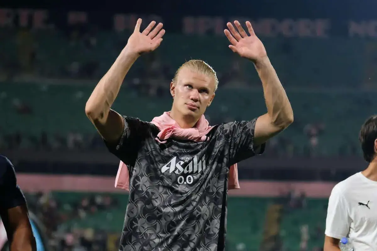 Erling Haaland z podniesionymi rękami, w koszulce z logo Asahi. Fani zastanawiają się nad jego datami dołączenia do drużyny.