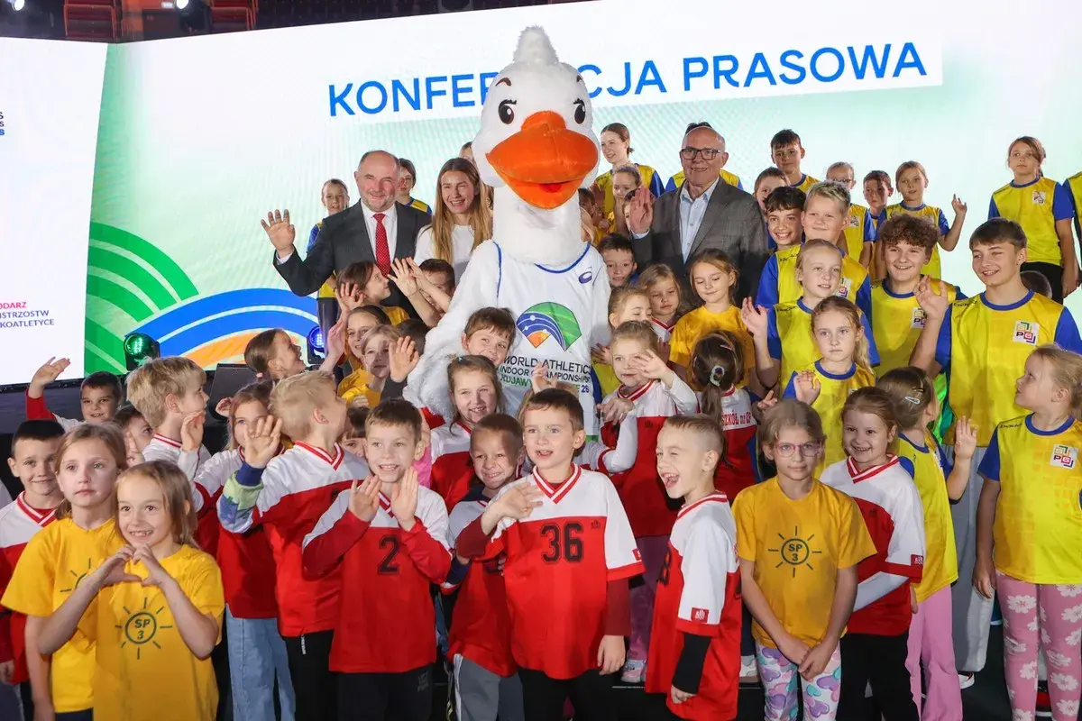 Uśmiechnięte dzieci i duża maskotka z logo lekkoatletycznych mistrzostw świata. Wszyscy pozują do zdjęcia, w tle napis "Konferencja prasowa".