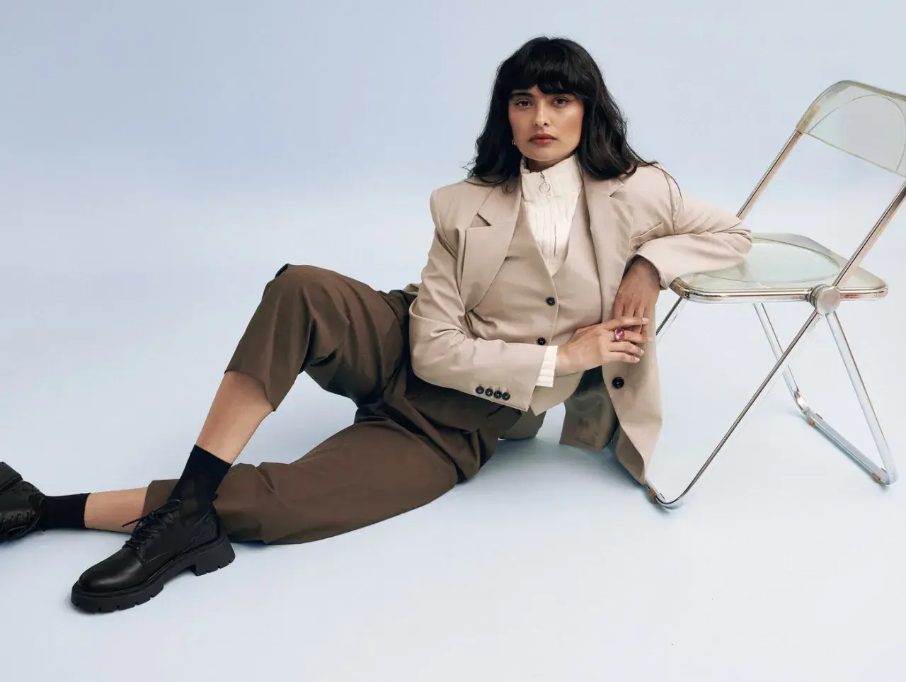 Femme en tenue professionnelle, blazer beige, pantalon marron, et chaussure entretien d'embauche femme noire. Assise près d'une chaise transparente.