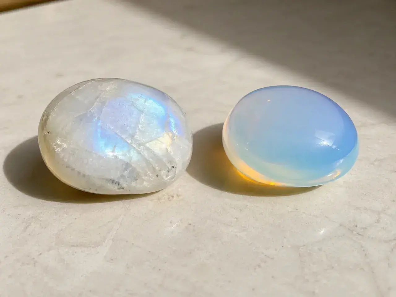 Dos piedras pulidas, una piedra luna iridiscente y otra de un azul lechoso, descansan sobre una superficie clara.