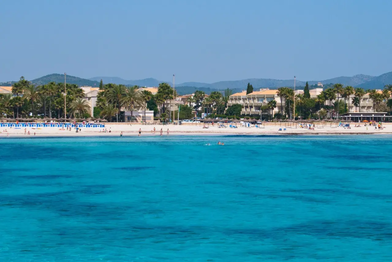 Türkisblaues Meer vor Sa Coma Altstadt mit Palmen und Hotels. Menschen genießen den Strand und das Wasser.