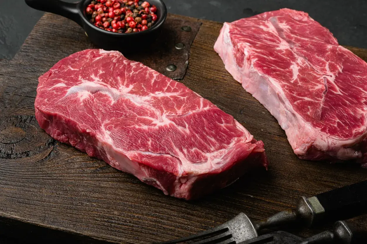 Deux beaux steaks de l angus beef, marbrés de gras, prêts à être cuisinés sur une planche en bois. Poivre rose en accompagnement.