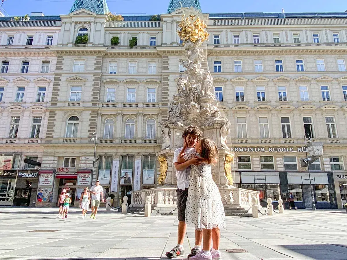 Due bambini si abbracciano davanti alla Colonna della Peste a Vienna. Altri bambini passeggiano sullo sfondo.