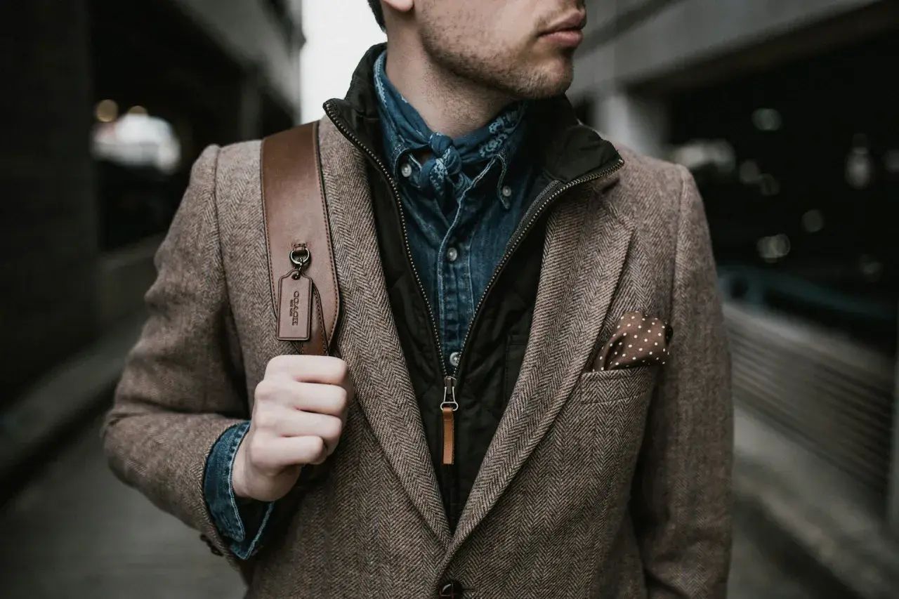 Mann im Tweed-Sakko mit Jeanshemd und Halstuch, perfekt für den party chic dress code male. Braune Ledertasche mit Coach-Anhänger.