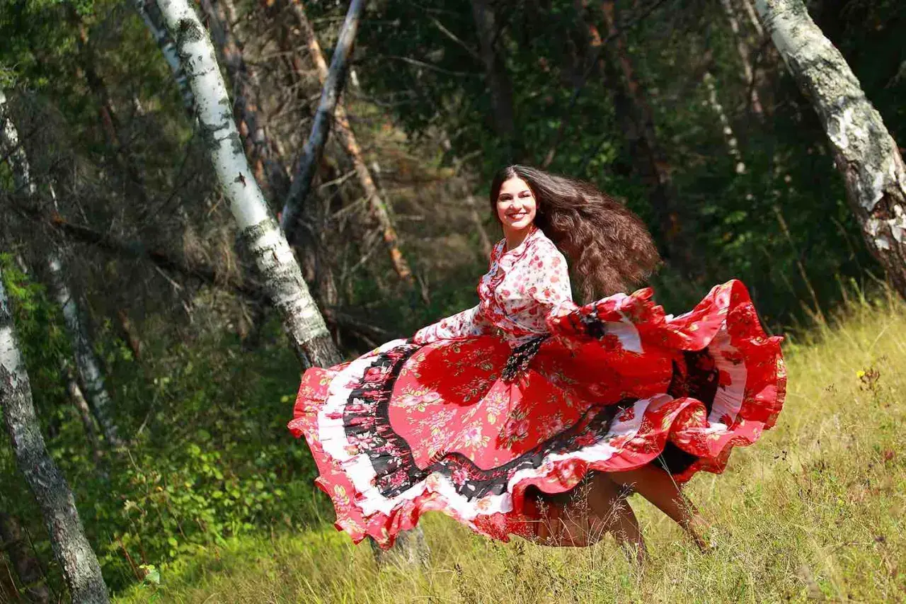 Una giovane donna con lunghi capelli scuri, vestita con un abito rosso svolazzante, danza in un bosco. Sembra una delle giostrai Sinti, piena di gioia e vitalità.