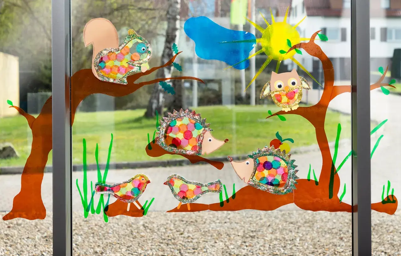 Bunte Fensterbilder basteln Kinder: Eichhörnchen, Igel, Eule & Vögel auf Ästen. Sonnenschein & Wolke im Hintergrund.