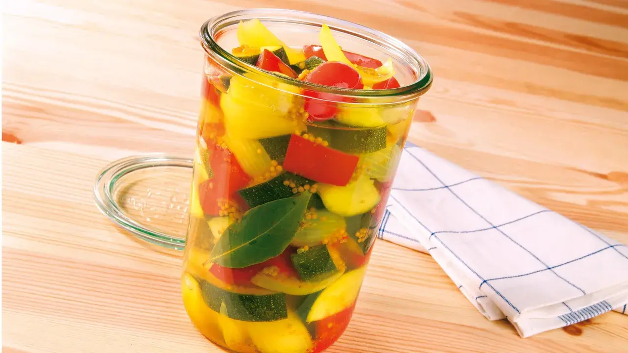 Ein Glas mit bunter Mischung aus Zucchini, Paprika und Tomaten, bereit zum Zucchini einkochen. Senfkörner und Lorbeerblätter sind ebenfalls sichtbar.