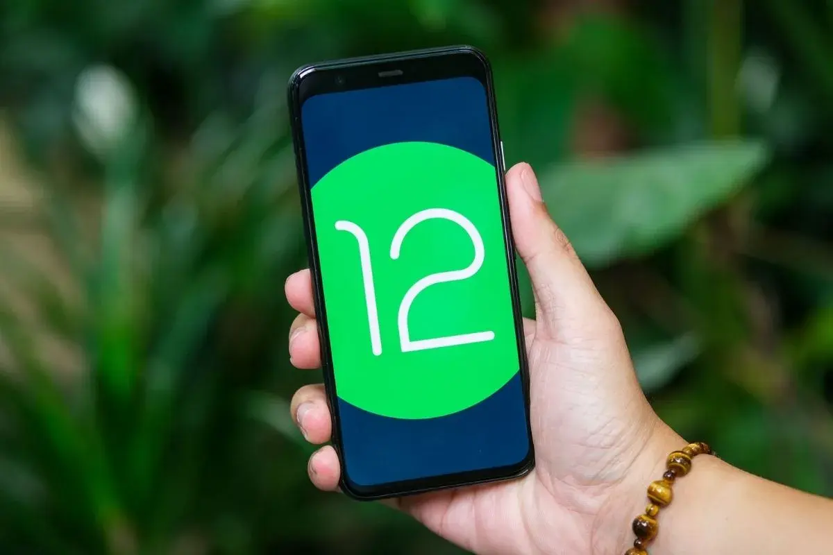 Dłoń trzyma smartfon z logo Android 12 na ekranie, na tle zieleni.