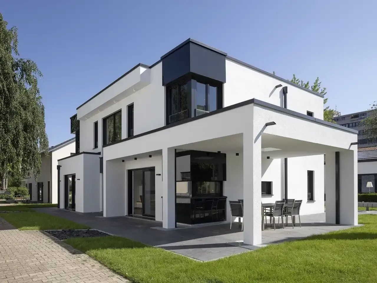 Ein modernes Haus mit Flachdach, weißen Wänden und schwarzen Fensterrahmen. Eine überdachte Terrasse mit Esstisch lädt zum Verweilen ein.