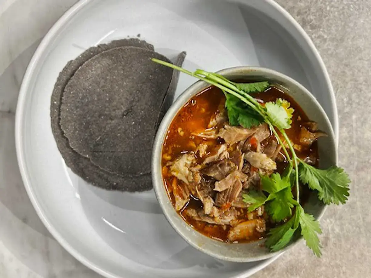 En skål med mustig el birria, toppad med koriander, serveras med mörka majstortillas på en vit tallrik.