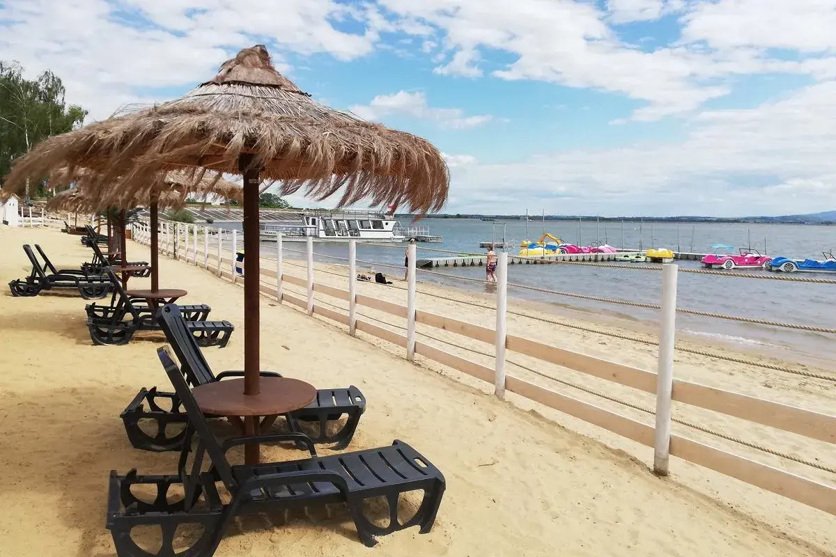 Plaża nad otmuchowskim jeziorem, leżaki pod strzechą, w oddali kolorowe rowerki wodne i statek wycieczkowy.