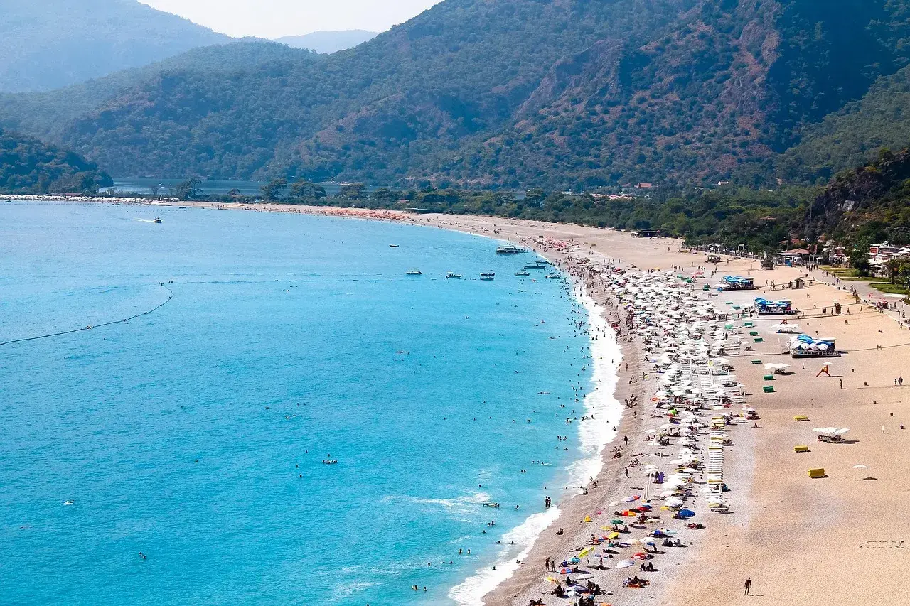 Tłum ludzi odpoczywa na piaszczystej plaży, kąpiąc się w turkusowym morzu. Widok na riwierę egejską z górzystym, zalesionym tłem.