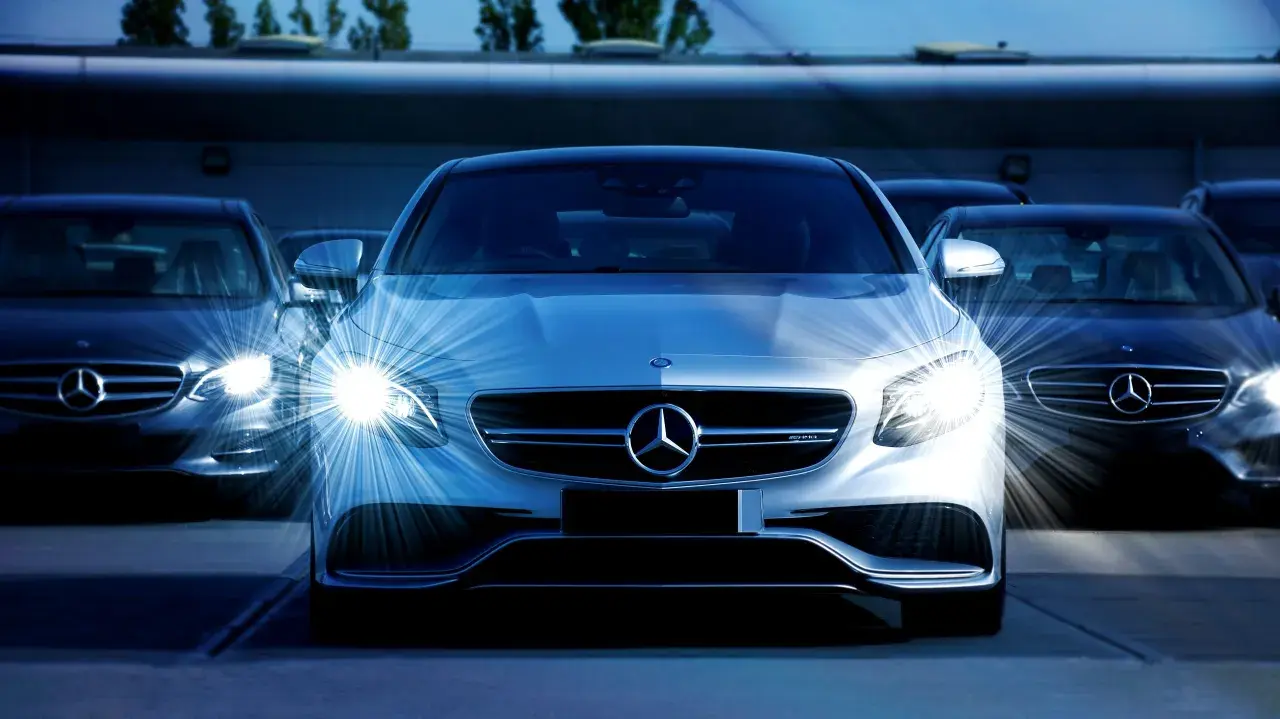 Biała hybryda plug-in Mercedes-Benz z włączonymi światłami, otoczona innymi ciemnymi samochodami tej marki.