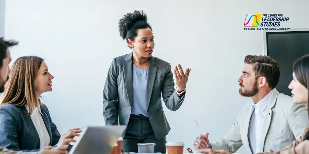 A diverse team engages in a communications leadership development program, with a woman in a grey suit leading the discussion.
