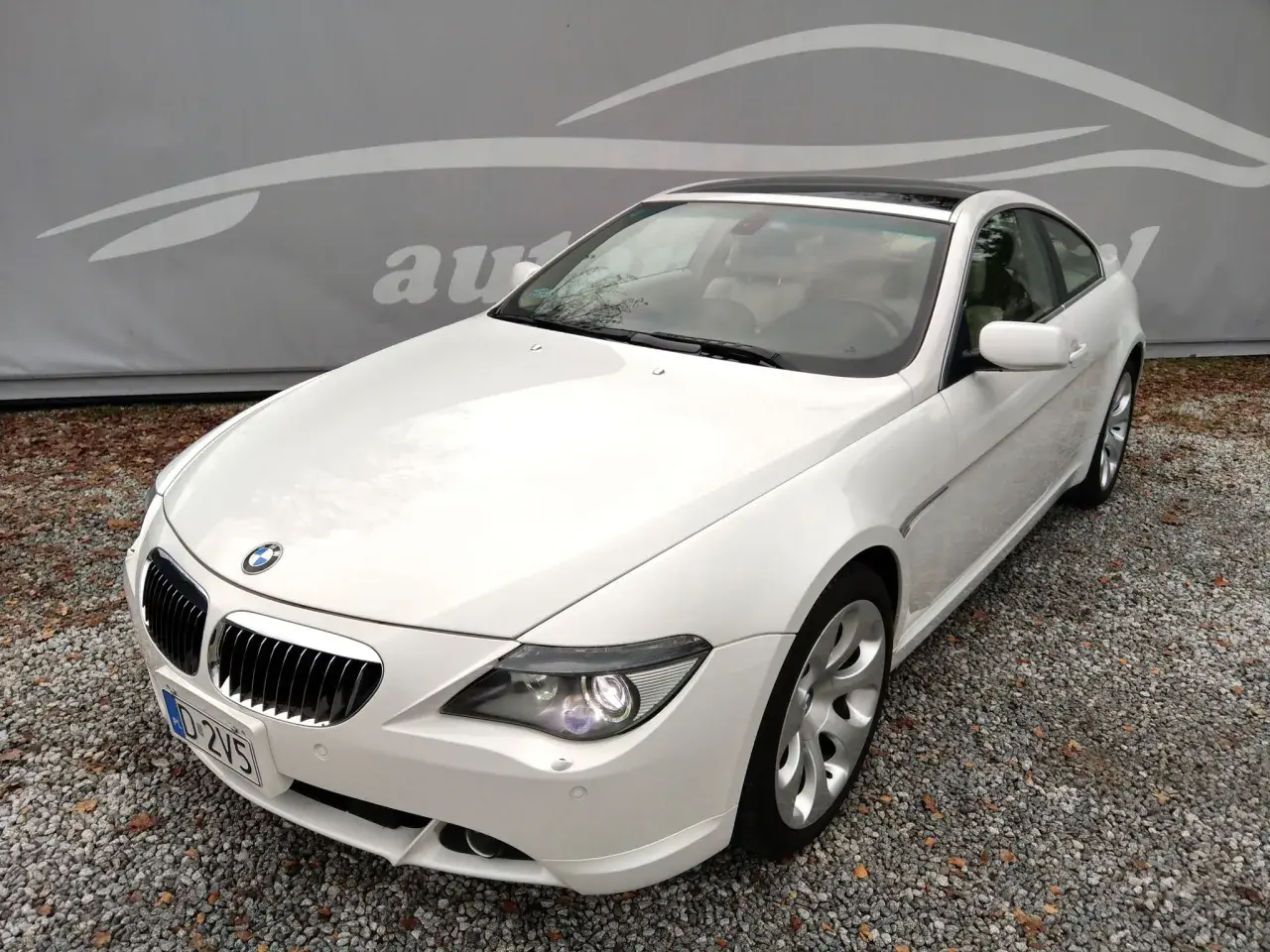 Białe BMW E63 coupe z jasnym wnętrzem, zaparkowane na żwirze, z logo autokomisu w tle.