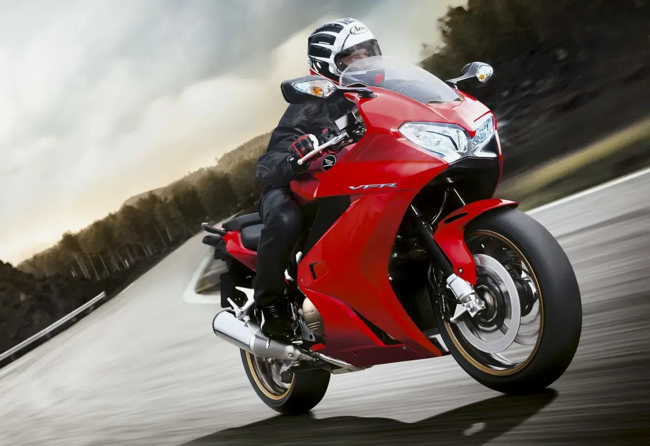 Motociclista en moto roja deportiva, con casco y equipo de cuero, inclinándose en una curva. El carenado de protección de la moto es aerodinámico y brillante.