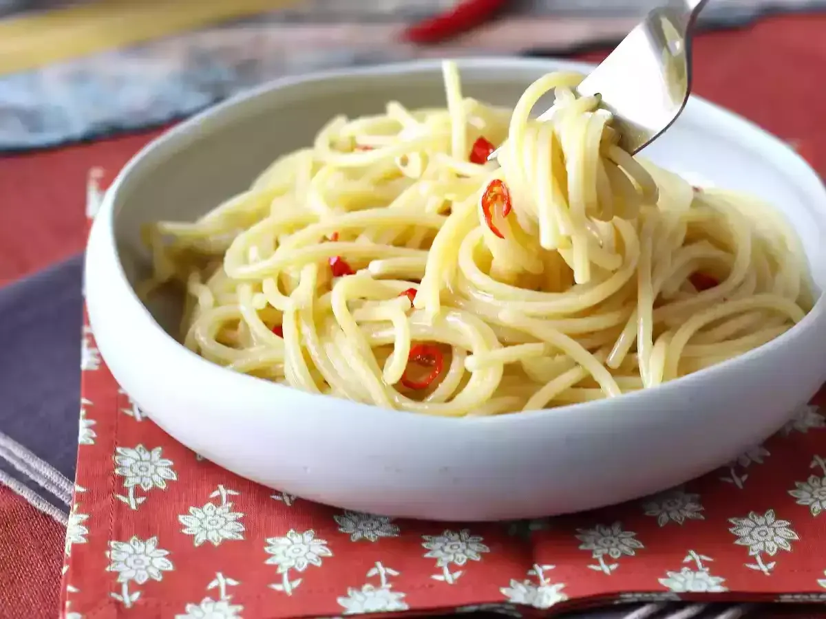 En skål med spaghetti aglio olio e peperoncino, där en gaffel lyfter upp en del av den oljiga pastan med små röda chilistrimlor.