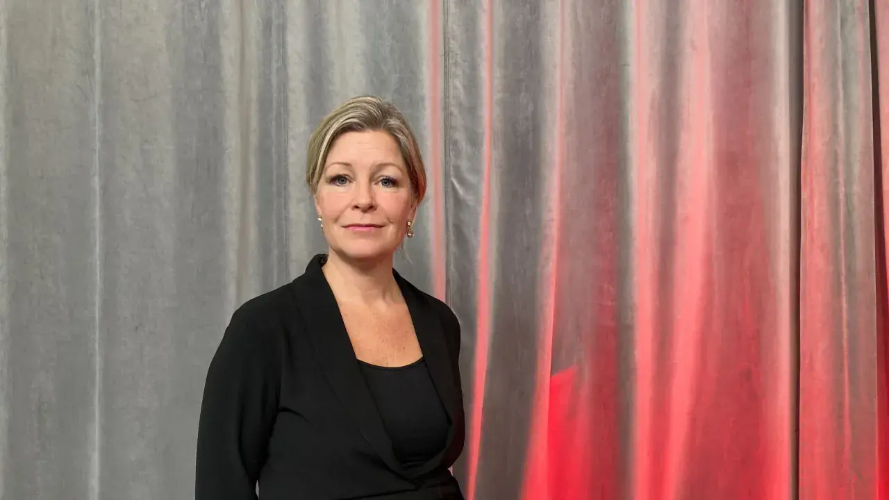 Johanna Fager Wettergren, en kvinna med blont hår och blå ögon, ler mot kameran framför en grå ridå med röd belysning.