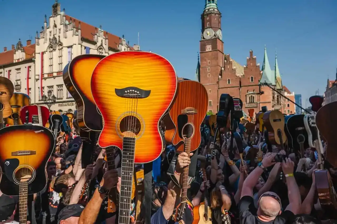 Tłum ludzi z gitarami wzniesionymi ku górze, świętujący gitarowy rekord świata na tle zabytkowej architektury.