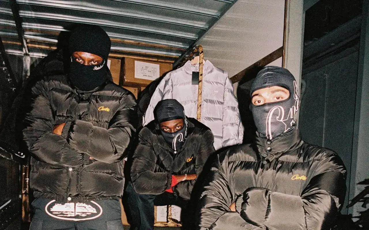 Drei Männer in schwarzen Daunenjacken mit dem **Corteiz logo** und Sturmhauben posieren vor einem Lagerhaus.