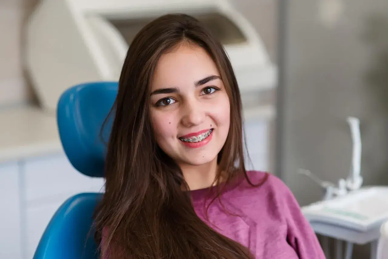 Uśmiechnięta nastolatka z kolorowym aparatem ruchomym na zębach siedzi na niebieskim fotelu dentystycznym.