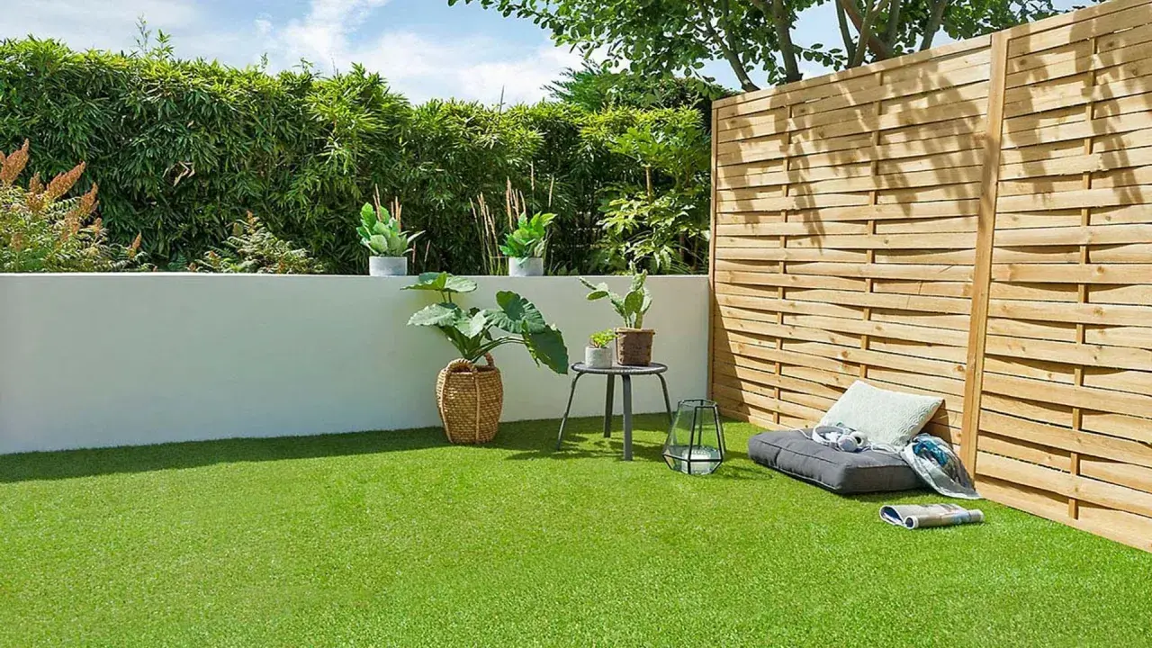 Un jardín moderno con césped artificial verde vibrante, rodeado de plantas y una valla de madera. Un rincón acogedor con cojines invita al descanso.