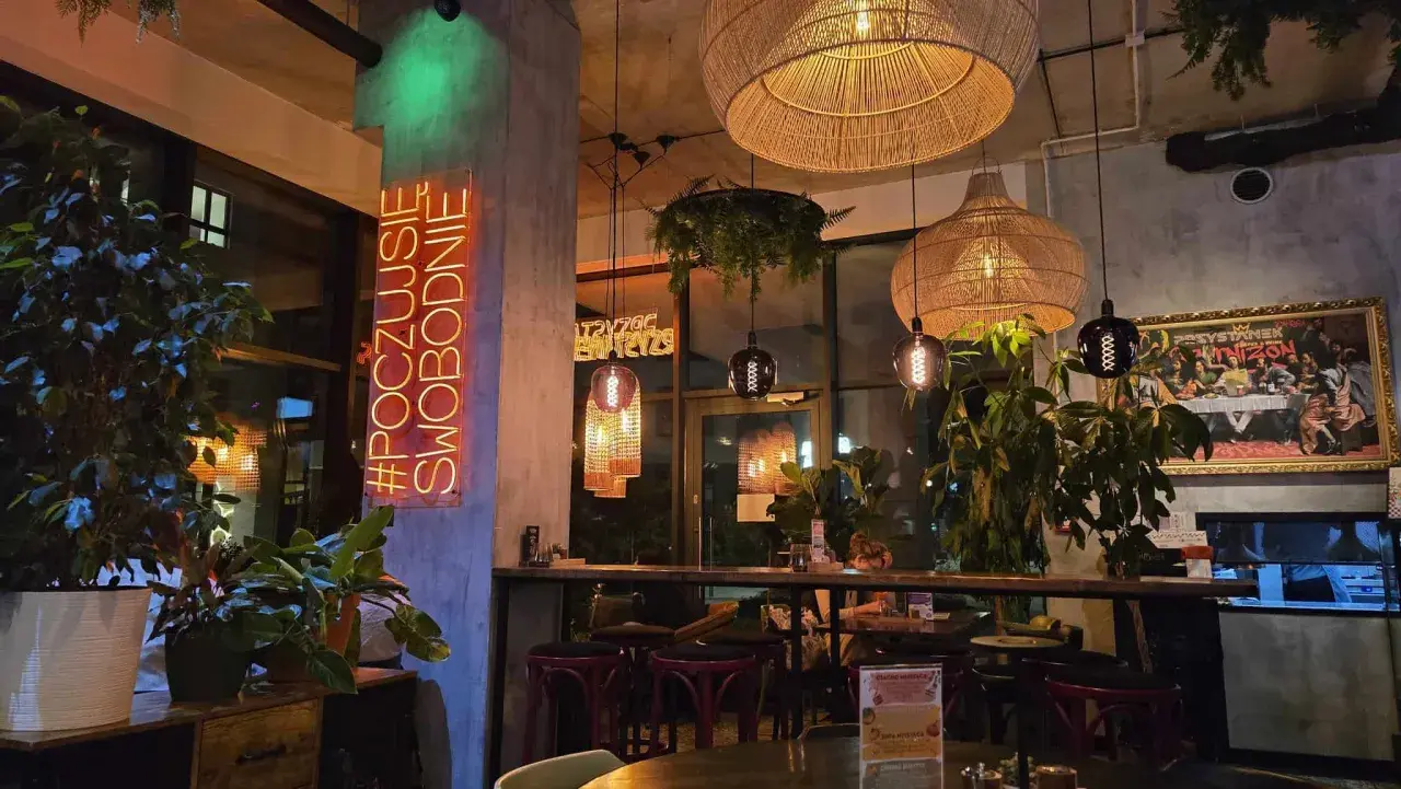 Wnętrze restauracji "Garnizon" z neonem "#POCZUJSIĘ SWOBODNIE", plecionymi lampami i roślinnością.
