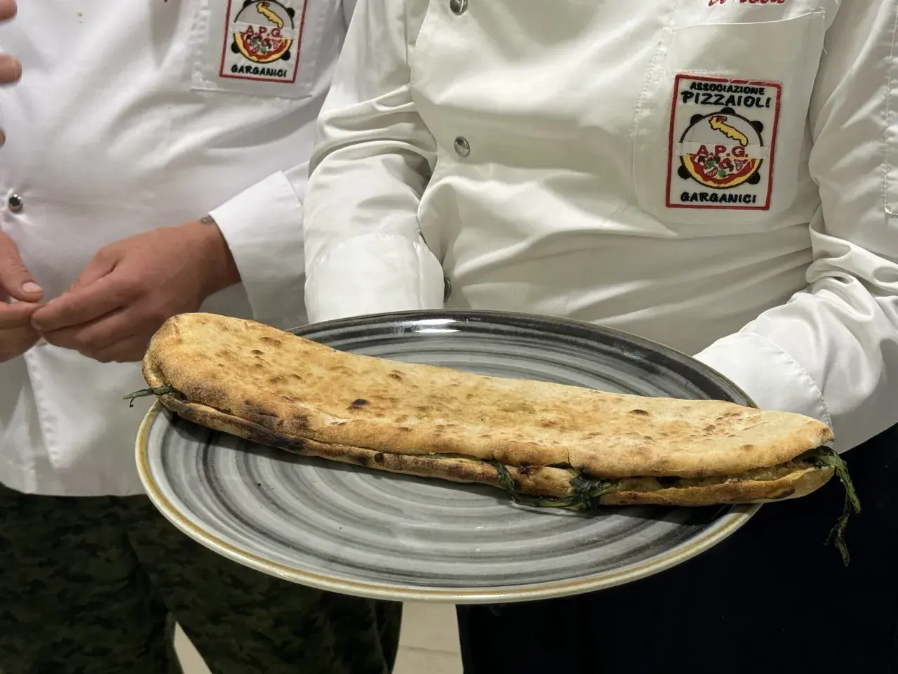 Una paposcia pugliese, ripiena e dorata, servita su un piatto grigio. Indossano giacche con stemmi dell'Associazione Pizzaioli Garganici.