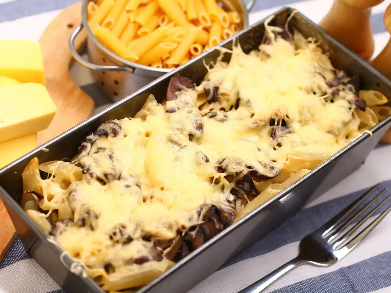 Pyszna zapiekanka z pieczarkami i makaronem penne, zapieczona pod złotym serem. Obok surowy makaron i kawałki sera.
