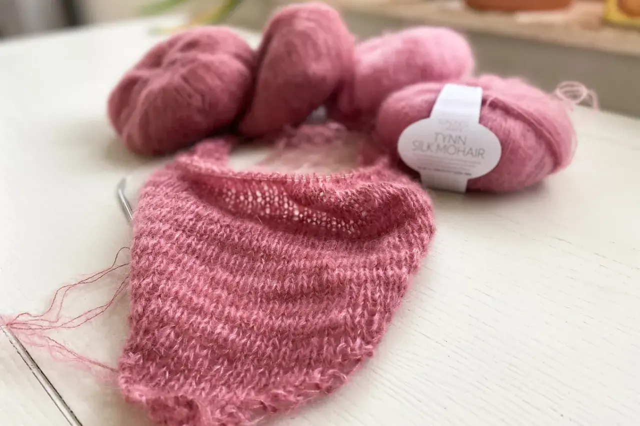 Zartrosa Mohair-Garn liegt neben einem Strickprojekt. Die feinen Fasern des Mohairs fusseln leicht und versprechen ein luftiges, weiches Ergebnis.