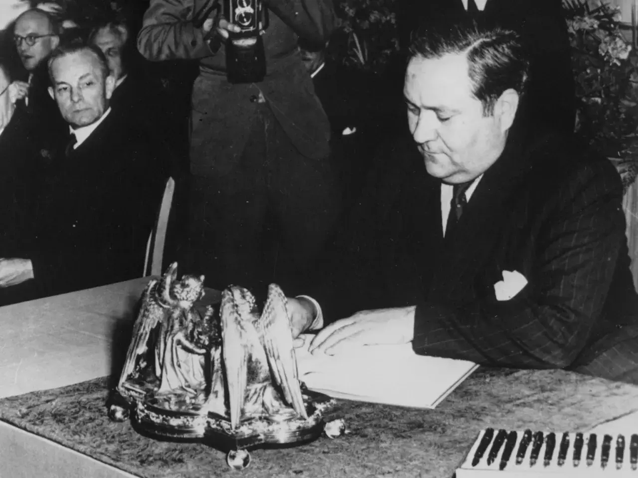 Carlo Schmid sitzt an einem Tisch und blickt auf ein Buch. Ein Fotograf hält eine Kamera.