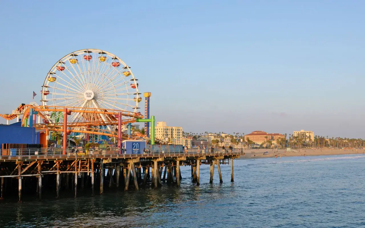 Der Santa Monica Pier mit seinem Riesenrad und Achterbahn ragt ins Meer. Im Hintergrund erstreckt sich der sonnige los angeles strand mit Palmen und Gebäuden.