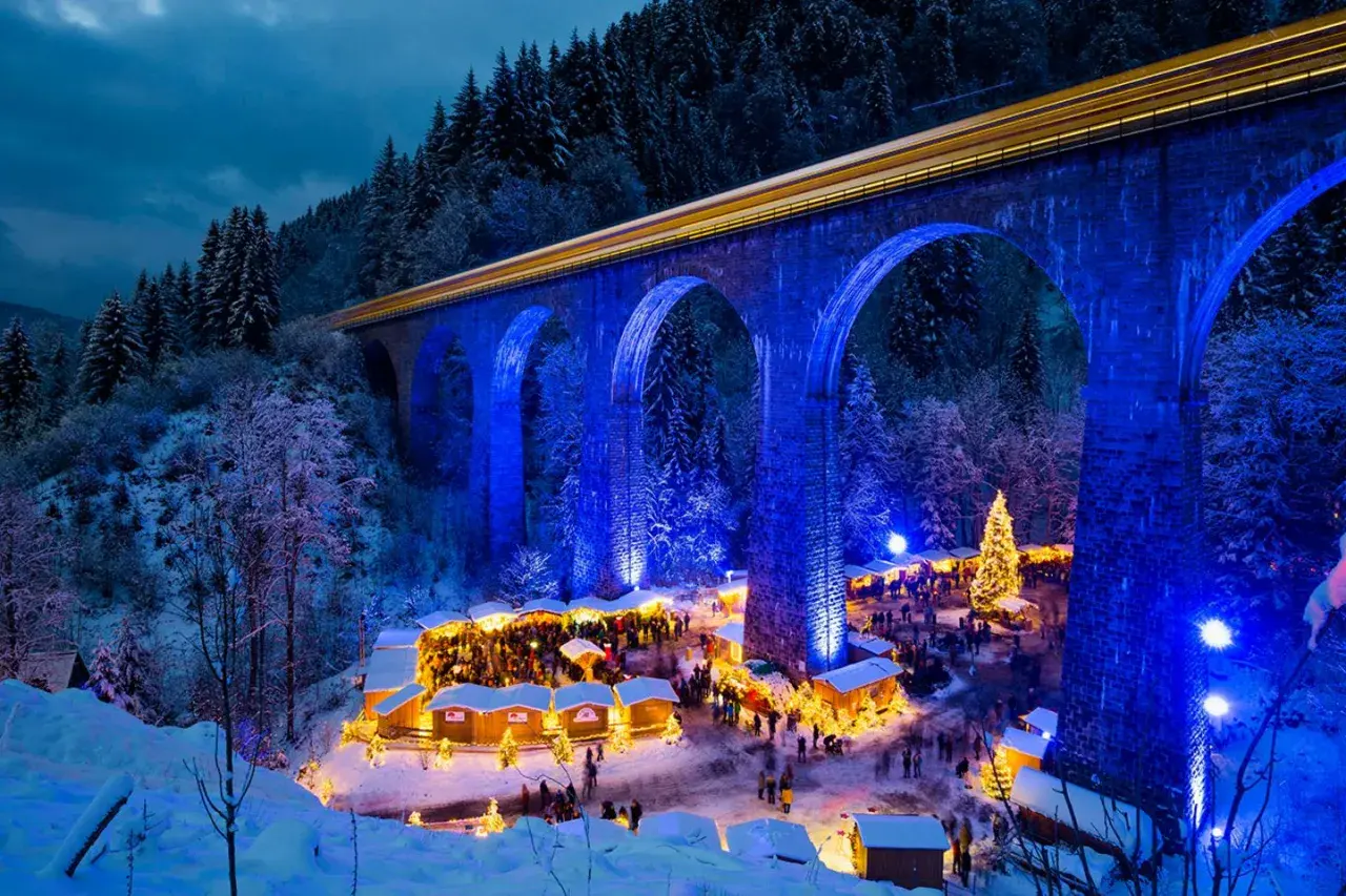 Magischer weihnachtsmarkt deutschland geheimtipp unter beleuchteter Brücke. Ein Zug fährt vorbei, während Besucher Buden und einen Weihnachtsbaum bestaunen.