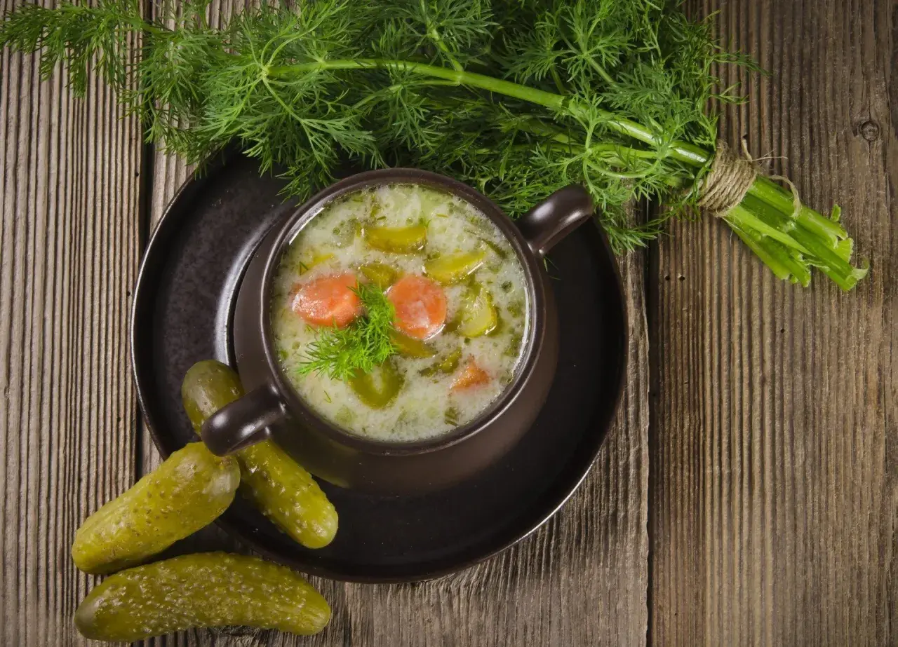 Aromaticzna zupa ogórkowa na żeberkach, podana w brązowej miseczce, ozdobiona koperkiem. Obok leżą chrupiące ogórki kiszone i pęczek świeżego koperku.