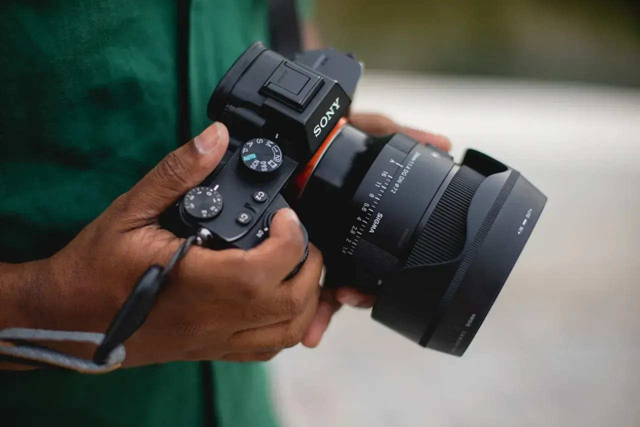 Dłonie trzymają aparat Sony z obiektywem Sigma. Widoczna jest skala ogniskowa obiektywu, wskazująca na jego możliwości.