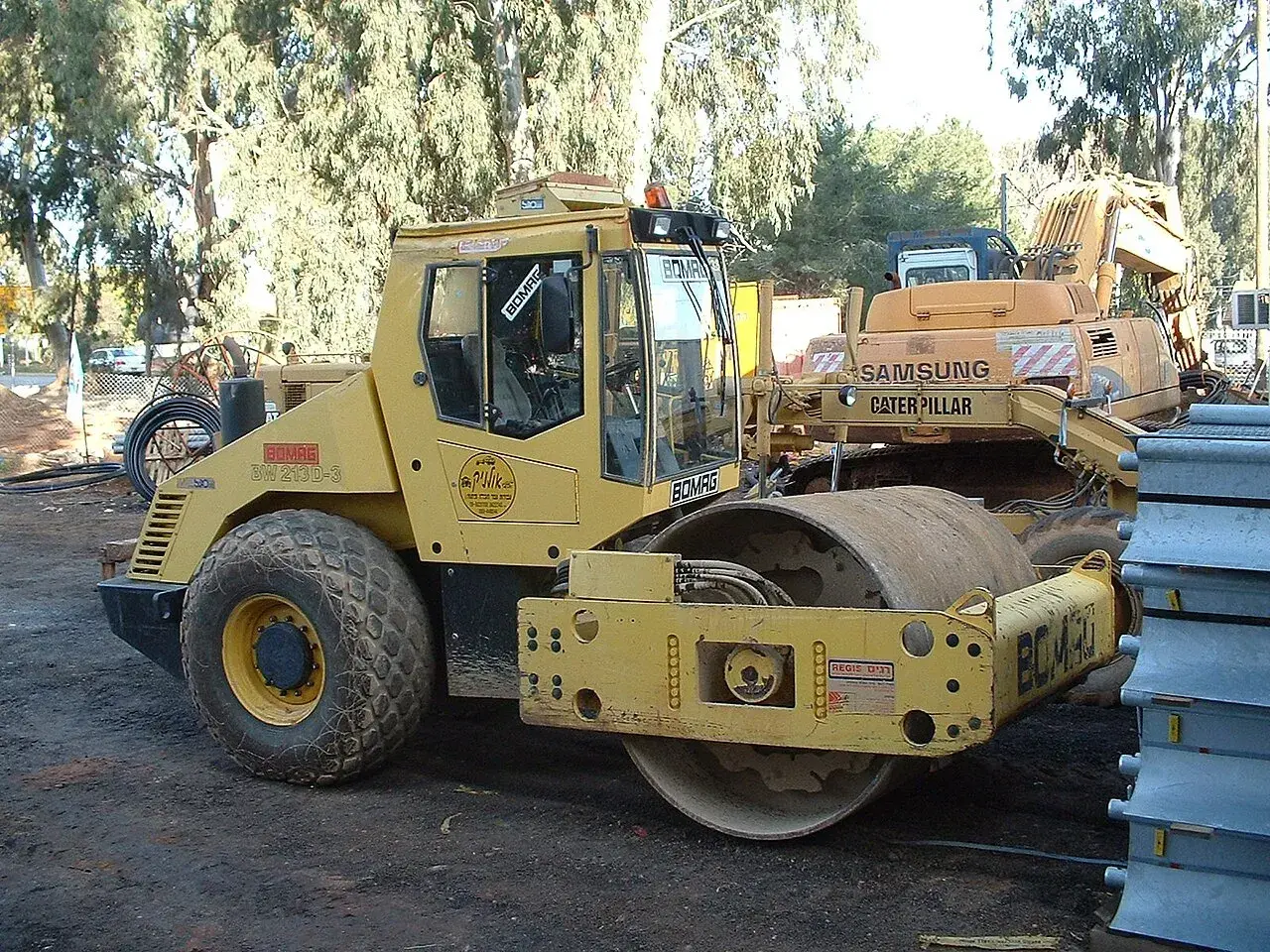 Żółty walec drogowy BOMAG BW 213D-3, obok koparka Samsung Caterpillar. Szerokość koparki nie jest widoczna, ale walec jest imponujący.