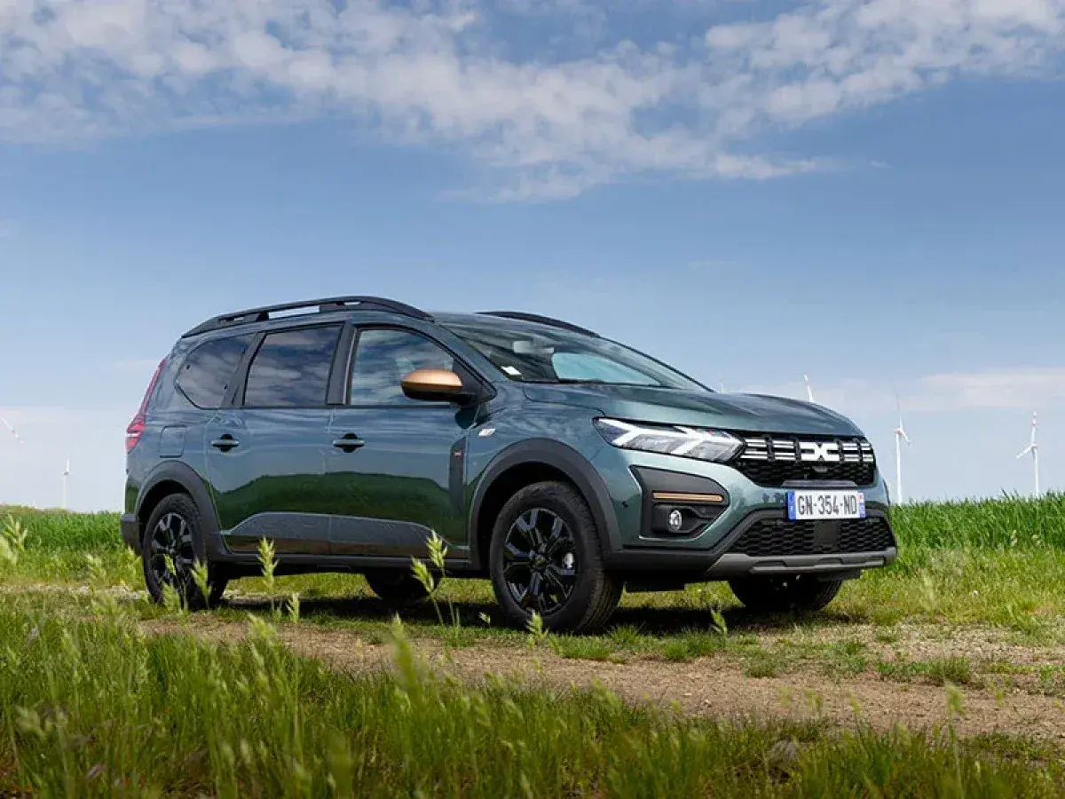 Un'auto familiare verde scuro, con barre sul tetto e dettagli color rame, parcheggiata su un sentiero sterrato in un campo con mulini a vento sullo sfondo.