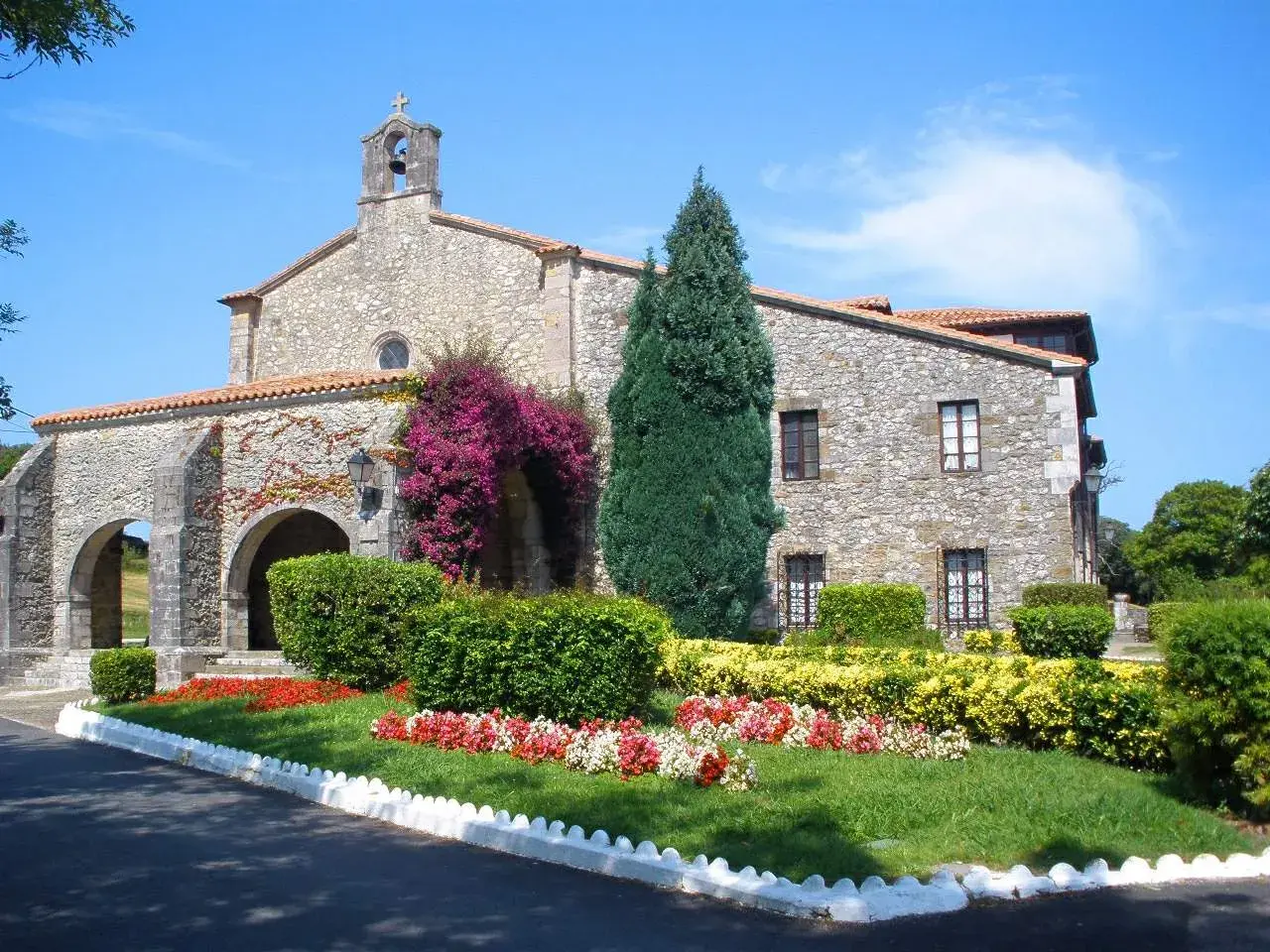 El **santuario de la Barquera**, con su fachada de piedra y arcos, se adorna con vibrantes buganvillas y un jardín florido bajo un cielo azul.