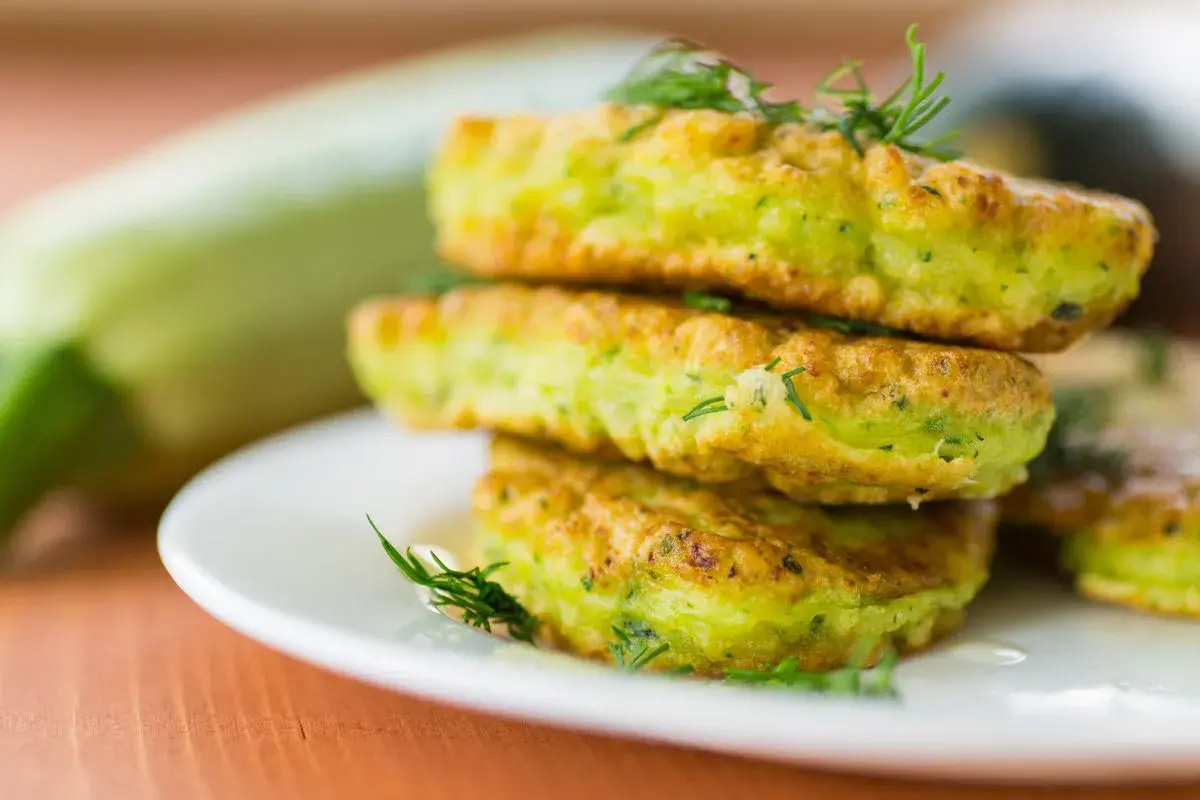 Frittelle di zucchine dorate e soffici, guarnite con aneto fresco, impilate su un piatto bianco. Sullo sfondo una zucchina intera.