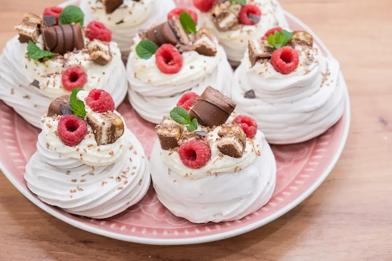 Słodkie mini pavlova z bitą śmietaną, malinami i kawałkami czekolady na różowym talerzu.