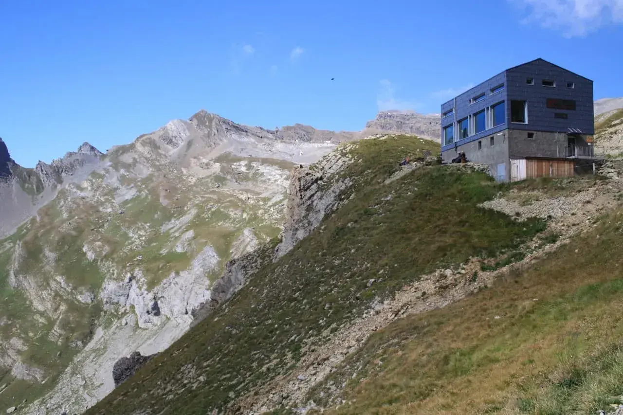 Moderne Hütte am Hang, Teil der Tour des Muverans, mit steilen Felswänden und grünen Bergwiesen im Hintergrund.