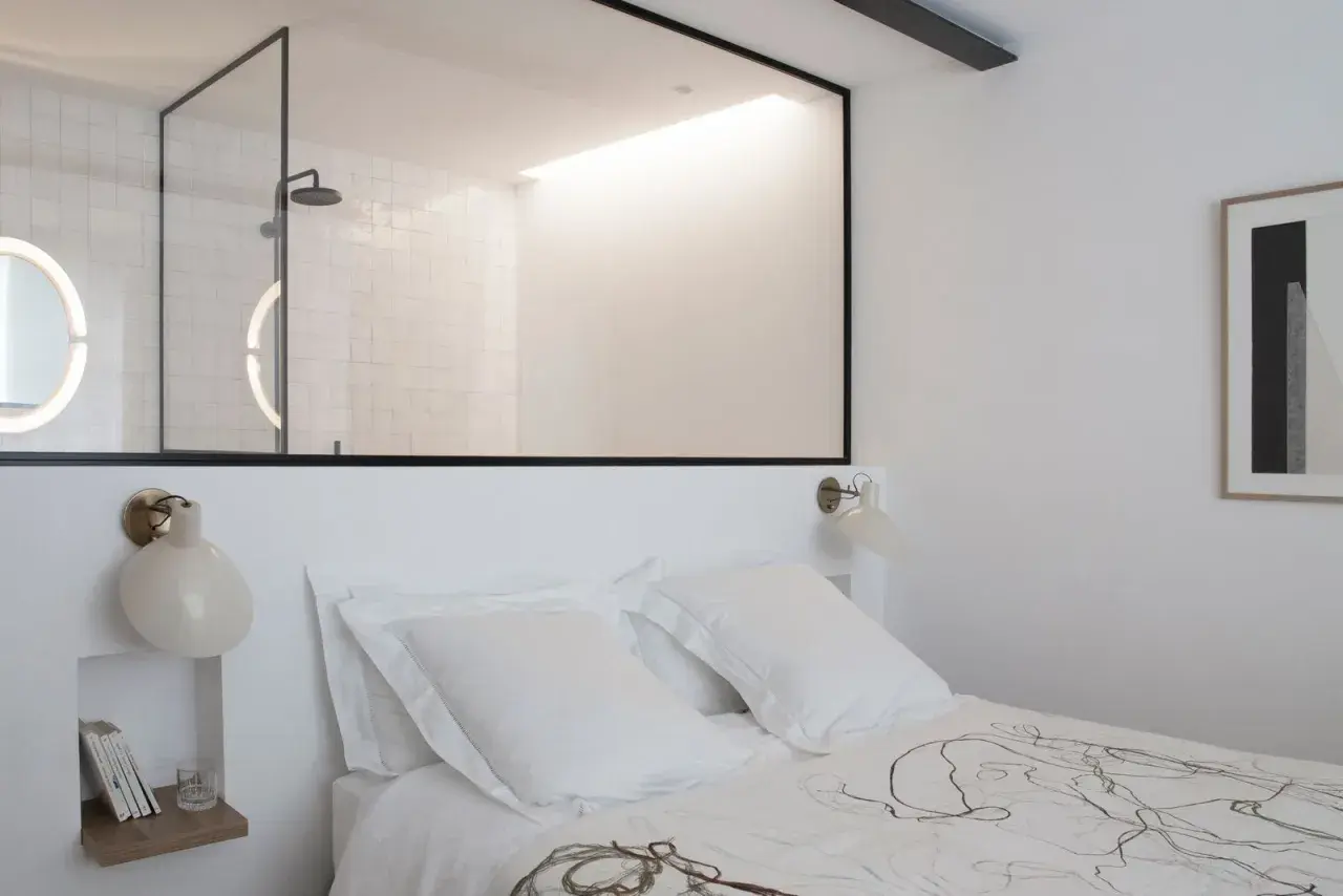 Stanza buia con letto bianco, una finestra tra due stanze rivela un bagno moderno con doccia e specchi luminosi.