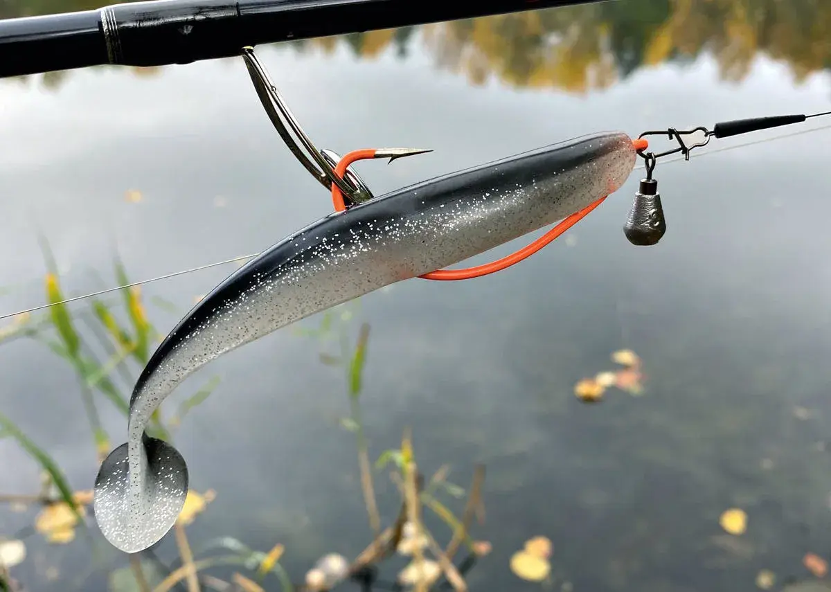 Zander-Montage: Ein Gummifisch mit Haken und Bleikopf hängt an einer Angelschnur über ruhigem Wasser.