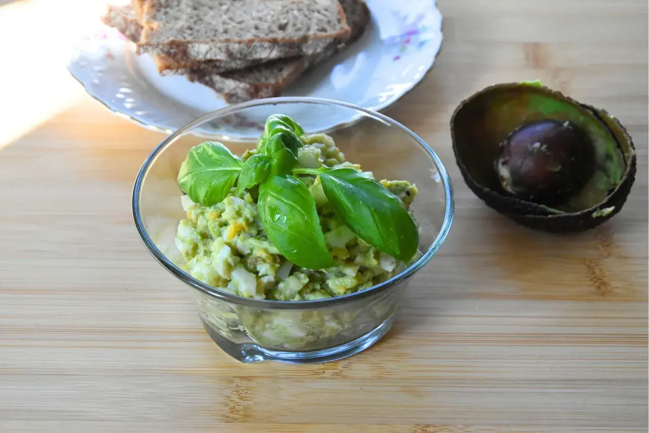 Pyszna pasta jajeczna z awokado, udekorowana bazylią, obok połówka awokado i kromki chleba.
