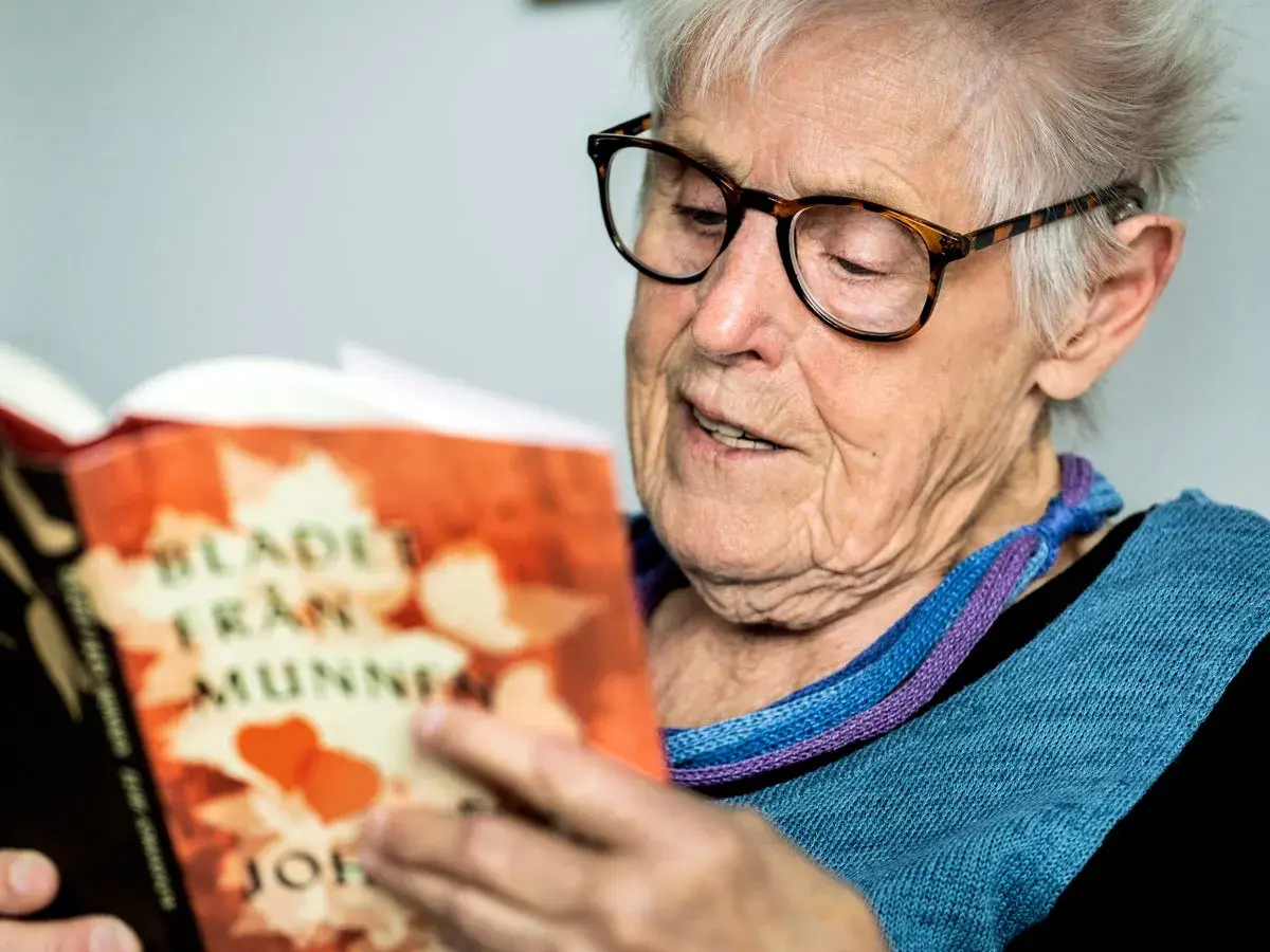 Äldre kvinna med glasögon läser en bok med titeln "Blad från munnen" av Elsie Johansson Thore.