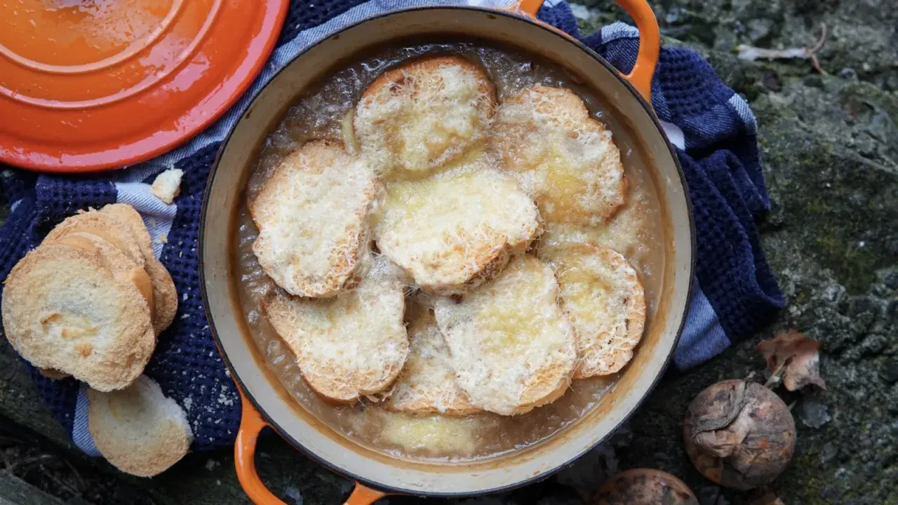 Zuppa di cipolle fumante in una casseruola arancione, con crostini dorati e formaggio fuso. Accanto, pane tostato e cipolle crude.