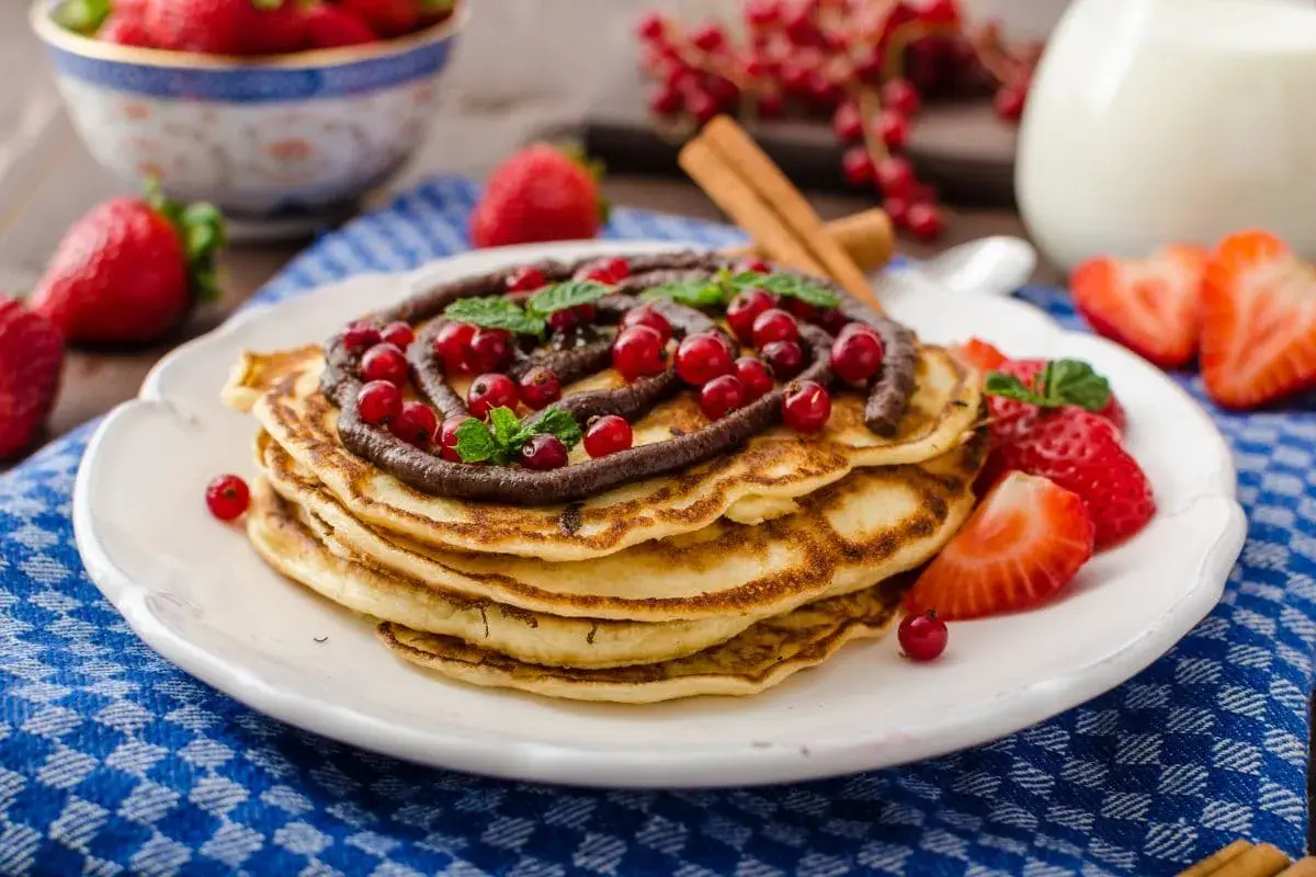 Stos puszystego ciasta na naleśniki z polewą czekoladową, porzeczkami i truskawkami.